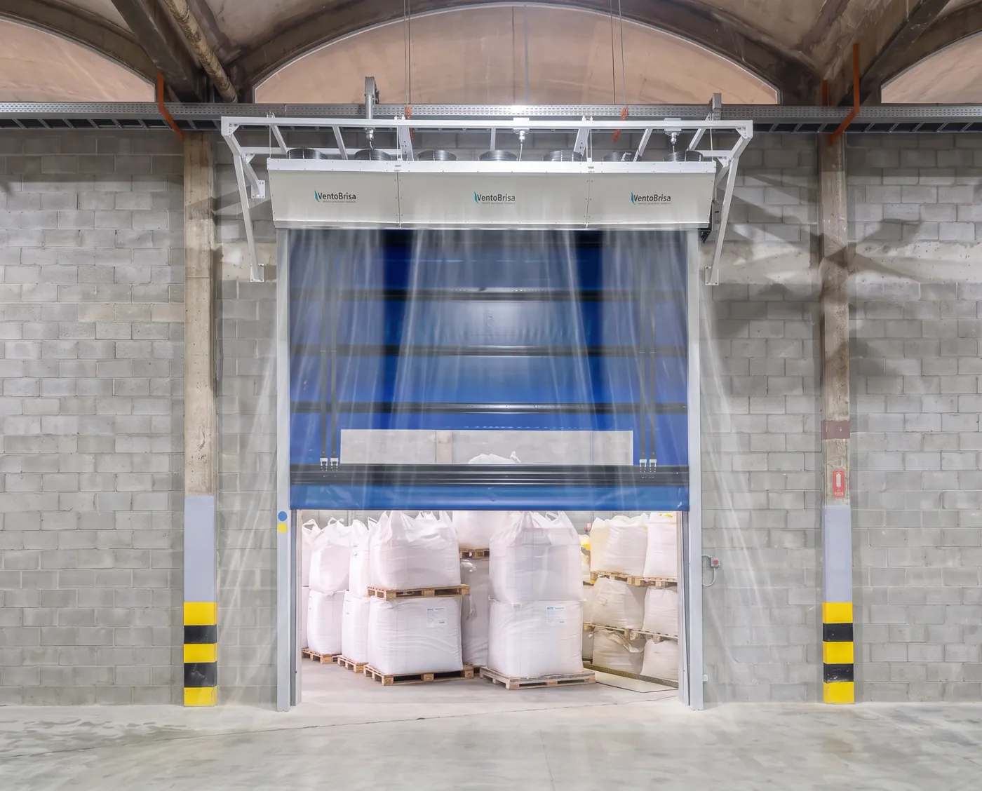 Cortina de ar industrial instalada sobre porta rápida azul aberta em armazém de big bags, com o fluxo de ar descendente ilustrado
